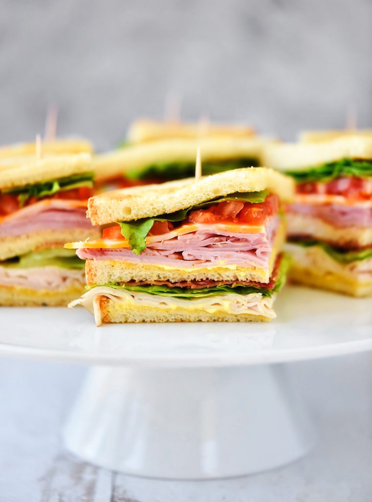 Club Sandwiches are stacked with ham, turkey, cheese, lettuce and tomato with a mustard and mayo mixture.