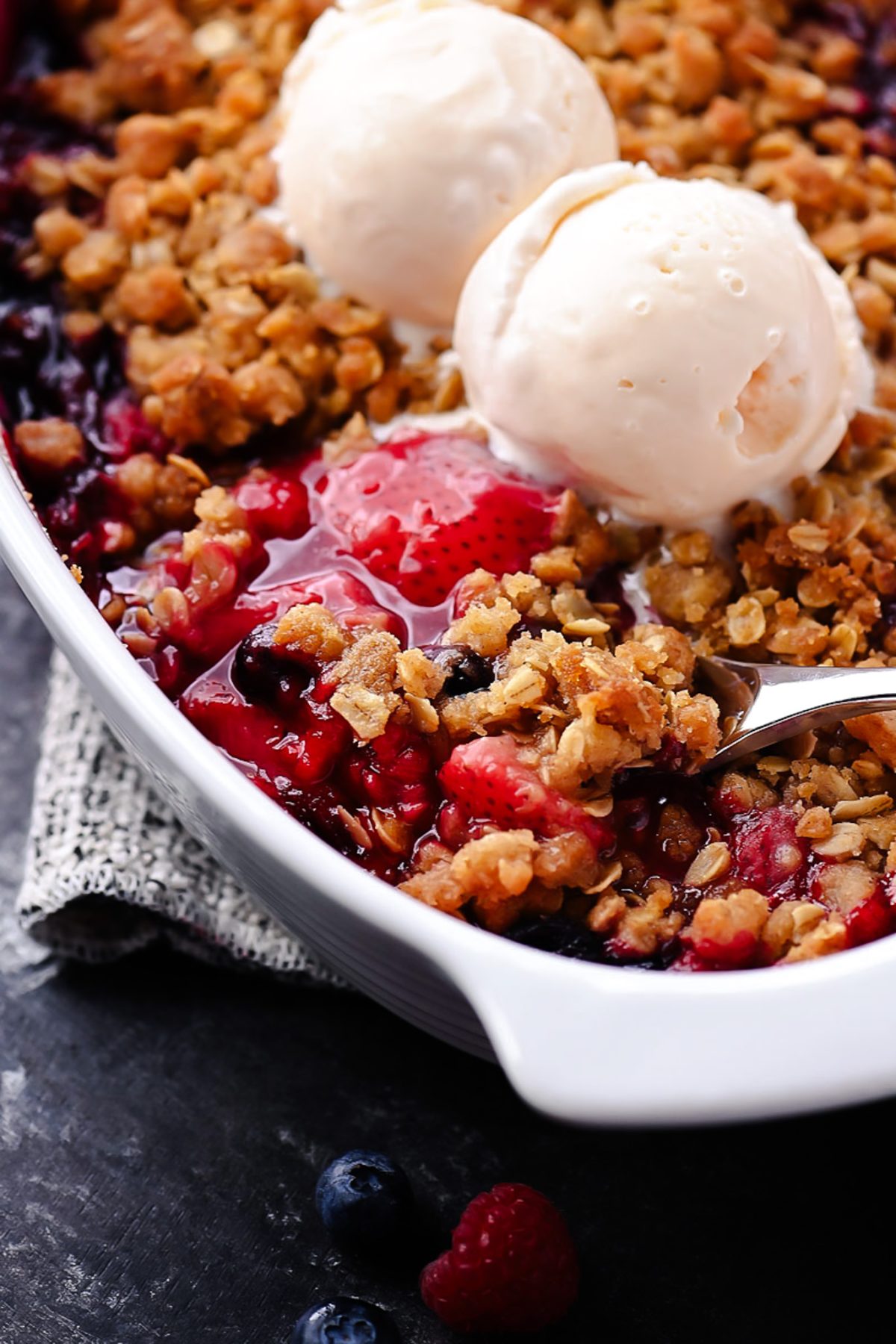 A warm berry crisp with bubbling mixed berries and a golden oat crumble topping, served in a white dish with a scoop of melting vanilla ice cream on top.