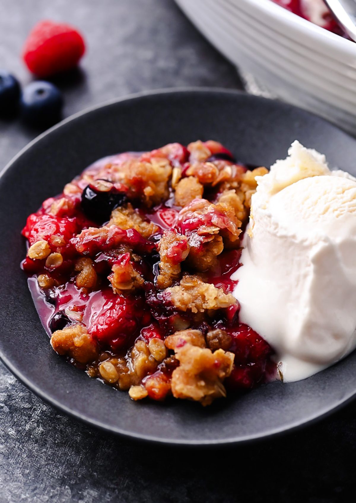 A warm berry crisp with bubbling mixed berries and a golden oat crumble topping, served in a white dish with a scoop of melting vanilla ice cream on top.