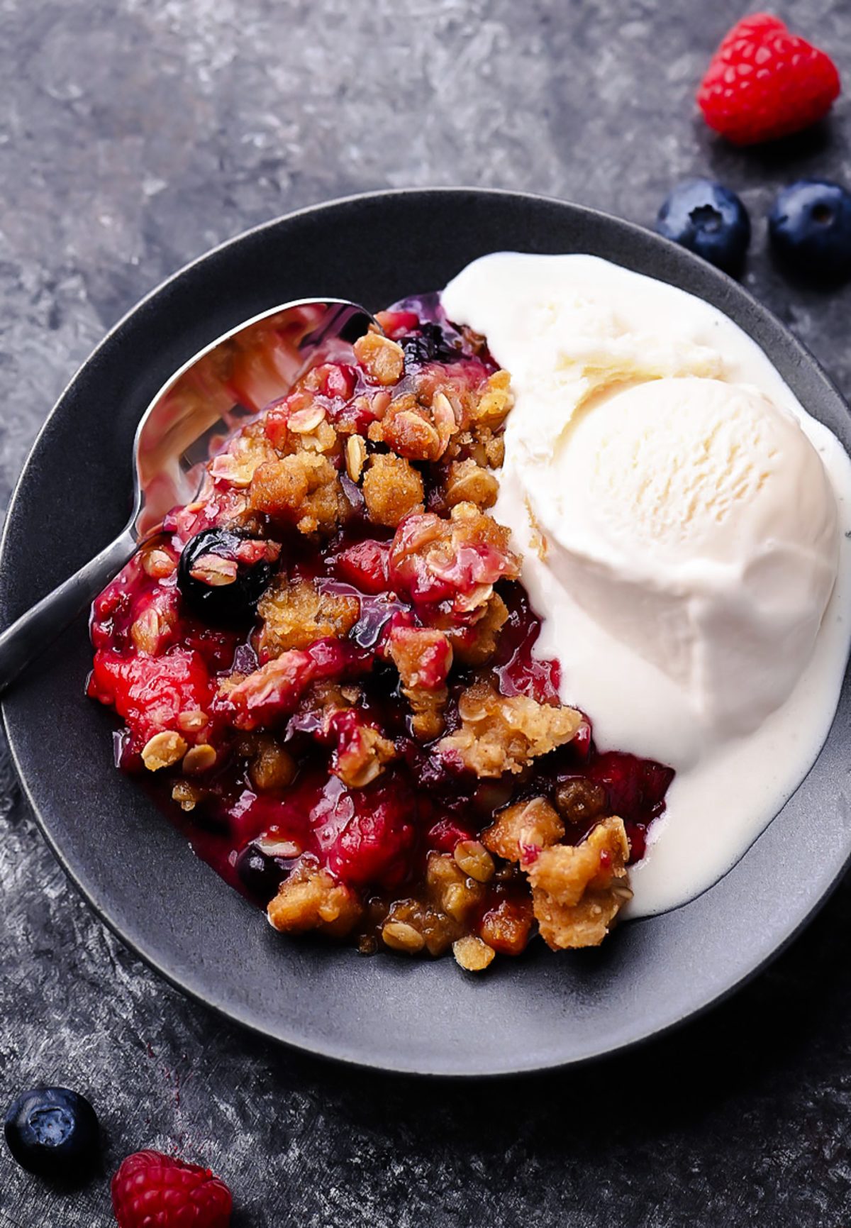 A warm berry crisp with bubbling mixed berries and a golden oat crumble topping, served in a white dish with a scoop of melting vanilla ice cream on top.