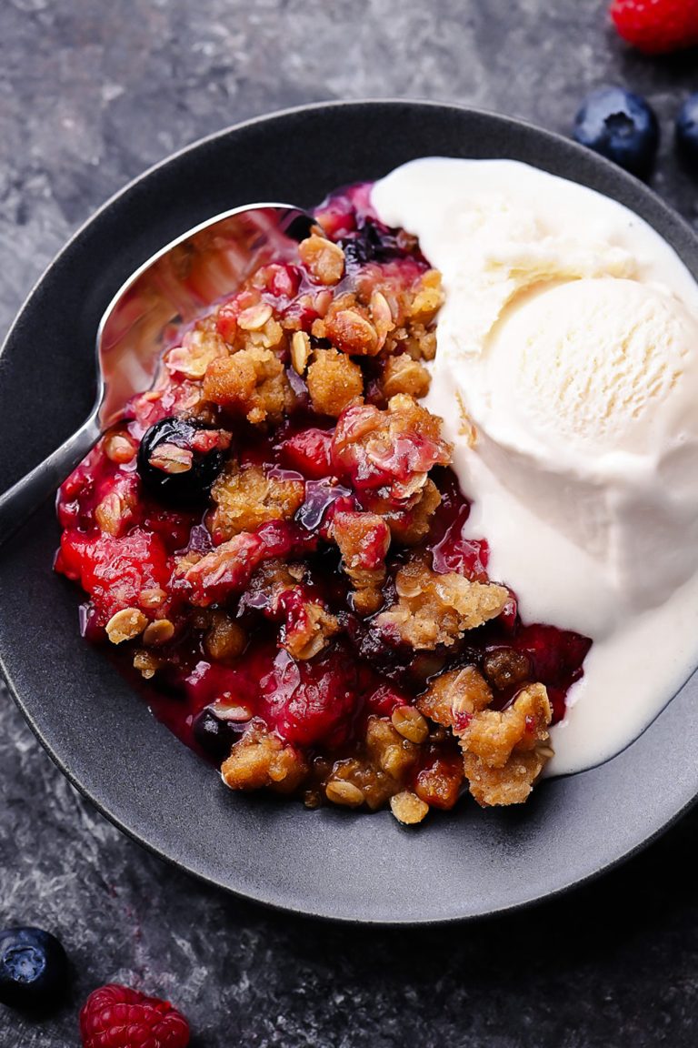 A warm berry crisp with bubbling mixed berries and a golden oat crumble topping, served in a white dish with a scoop of melting vanilla ice cream on top.