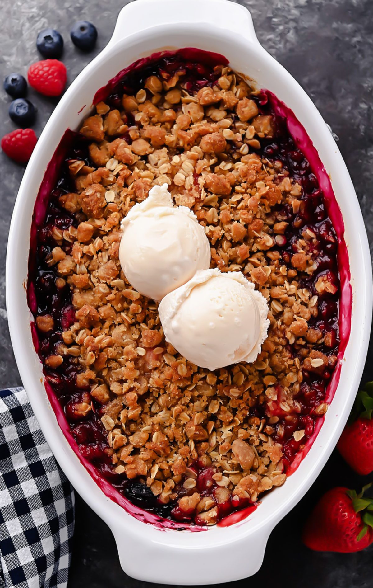 A warm berry crisp with bubbling mixed berries and a golden oat crumble topping, served in a white dish with a scoop of melting vanilla ice cream on top.